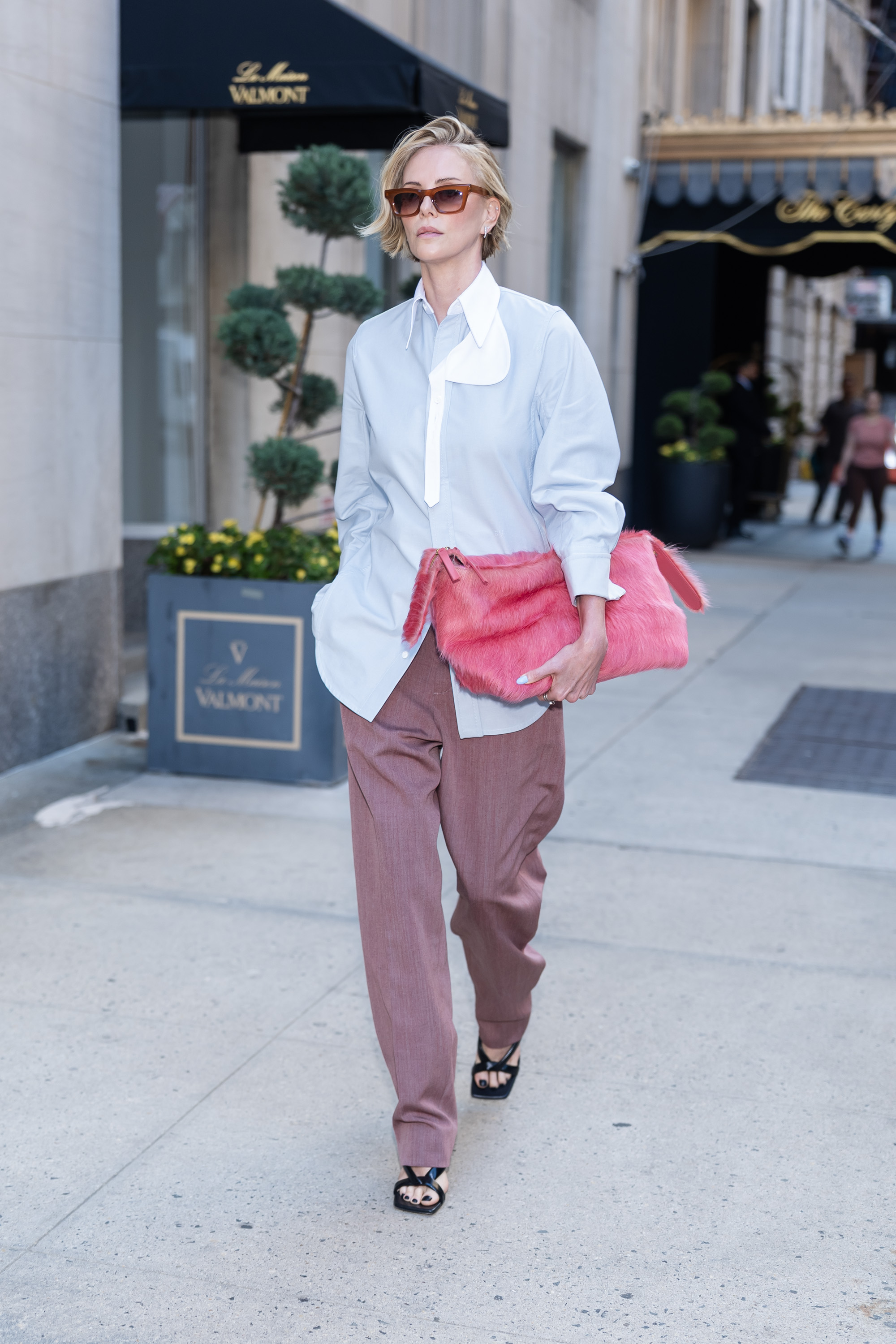 Charlize Theron walks down the street outside wearing mauve trousers with a baby blue shirt and a pink clutch bag.