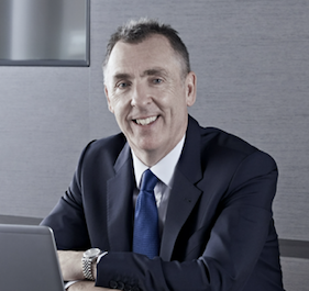 Headshot of a man in a dark grey suit and blue tie seated in front of a lap top