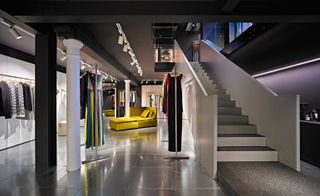 A clothing store with clothing displayed on rails, a yellow sofa, a white staircase and white spot lights above.