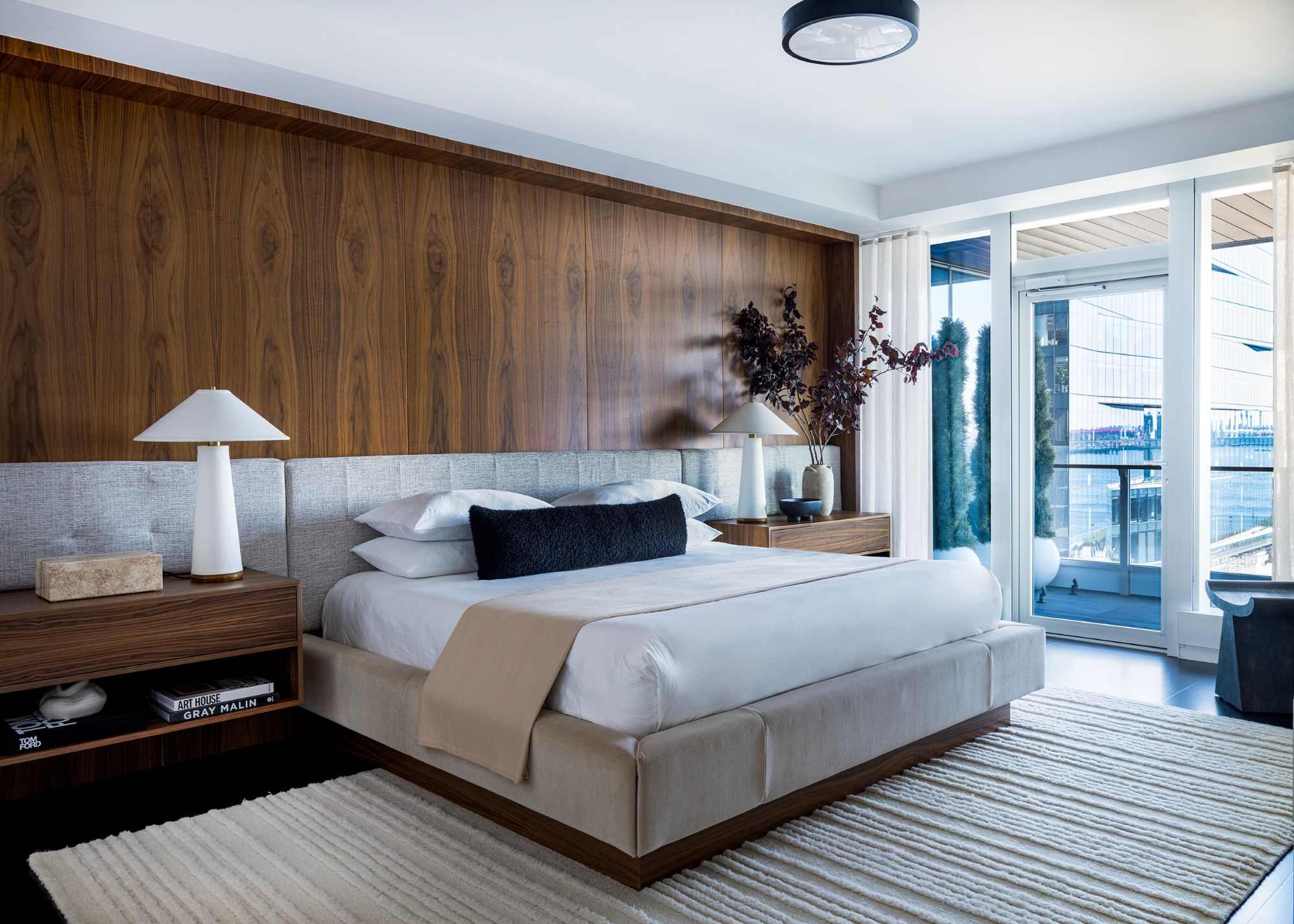 A large modern apartment bedroom with a wood paneled wall above the headboard, a large king-sized bed with white bedding, and a beige textured rug on the floor.