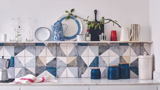 A white kitchen unit with drawers and shelf with a splashback of geometric patterned white, blue and grey, blue chair and marbled worktop, and blue and white patterned light shade.