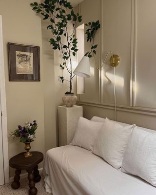 Neutral corner of living room with white couch, gilded sconce lighting, plant in a ceramic vase on stool in the corner and sculpted wooden small stool in view underneath small frame