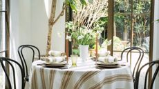 round table with striped tablecloth, four black bentwood chairs, place settings with plates, placemats, and tall taper candlesticks, and a centre vase with flowers