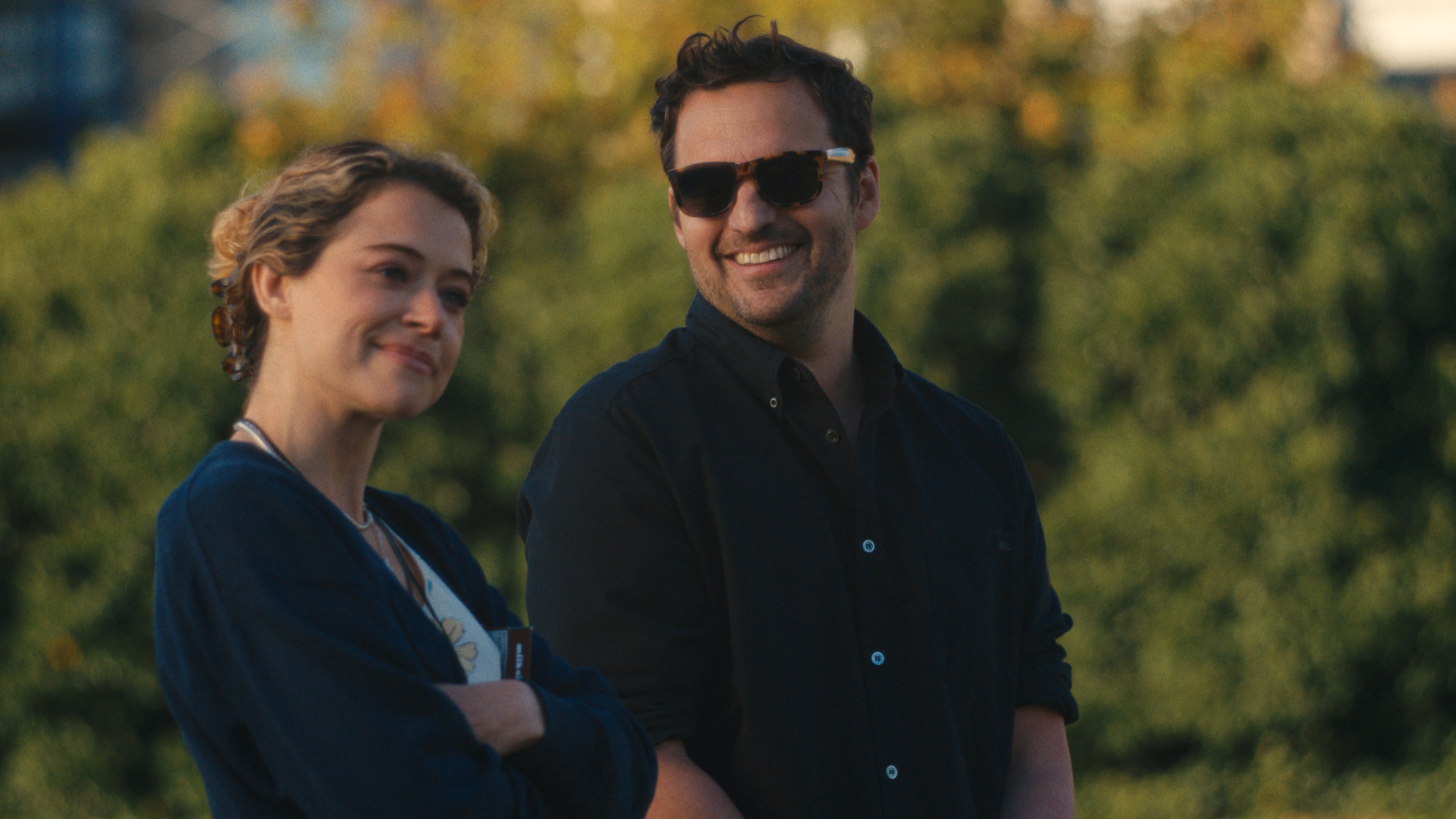 Karl (Jake Johnson) wearing sunglasses and smiling at Paula (Tatiana Maslany) who is smirking at something off camera with her arms crossed.