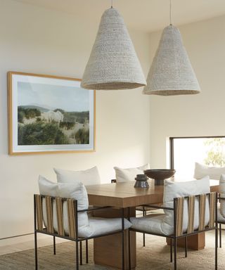 Relaxed dining room with cream painted walls, two large hanging pendants in natural, textured cream material, wooden rectangular dining table with dining chairs with metal frames, large webbed backing and cream upholstered seat and back cushions, rug below dining table, painting on wall, decorative vases and bowls on table