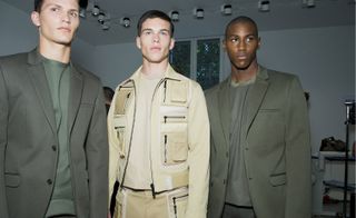 Three models wearing Calvin Klein clothing, two in olive colored suits and one wearing khaki colored suit