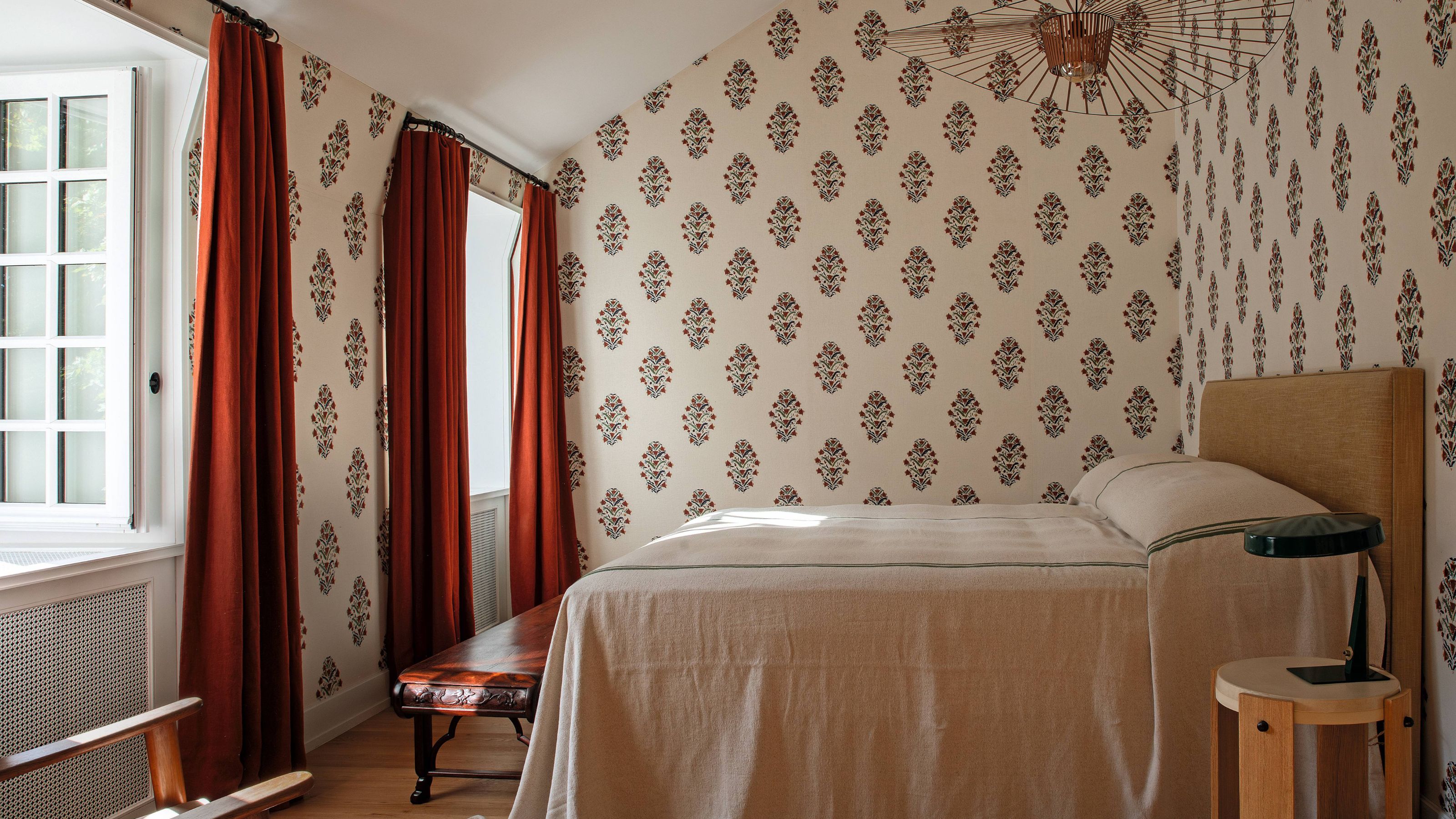 a block print floral bedroom with sloped ceiling, dark red drapes and a relaxed bed styled with traditional vintage french linen