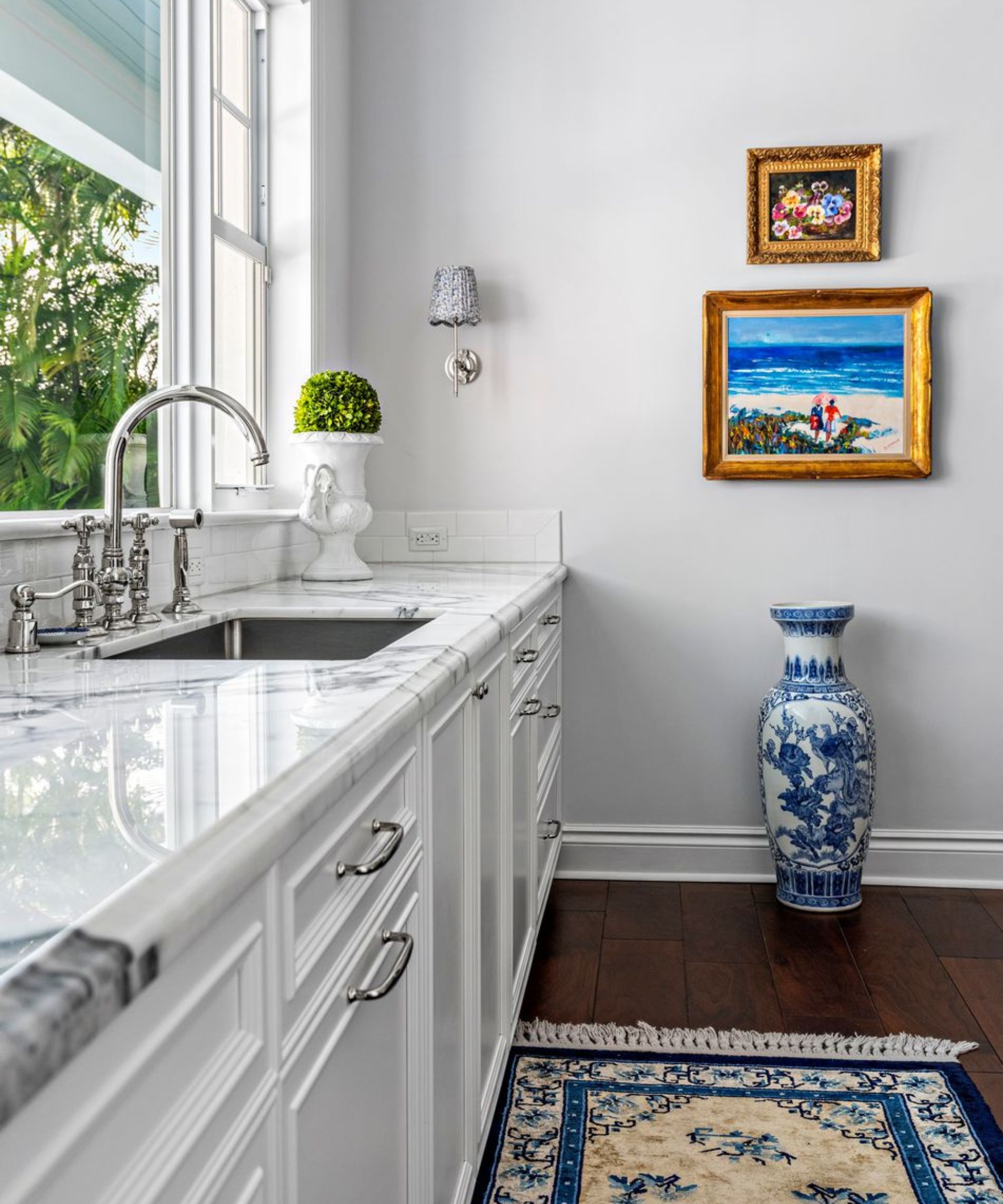 Kitchen with marble countertops, white cabinets, large window, wood floor and blue and white rug, large antique vase and ornate picture frames with little paintings on wall