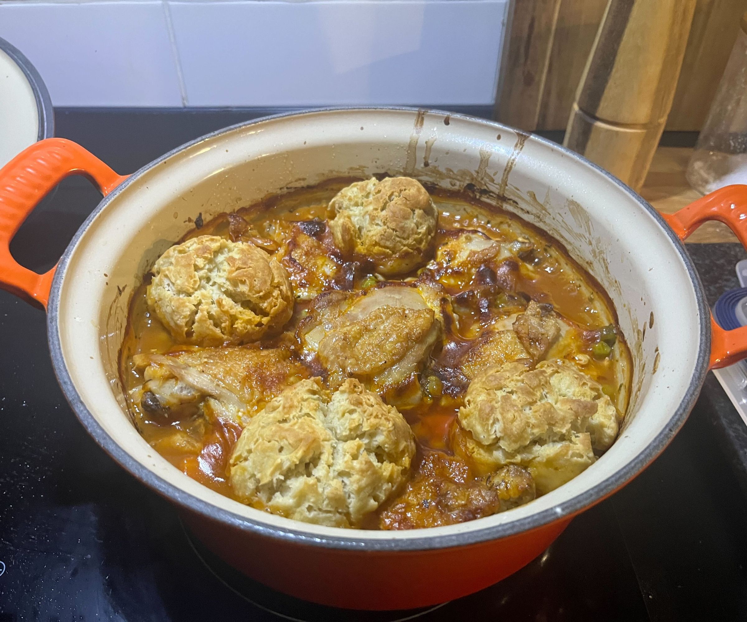 A golden brown chicken casserole with crispy dumplings in an orange cast iron Dutch oven, on a black stovetop