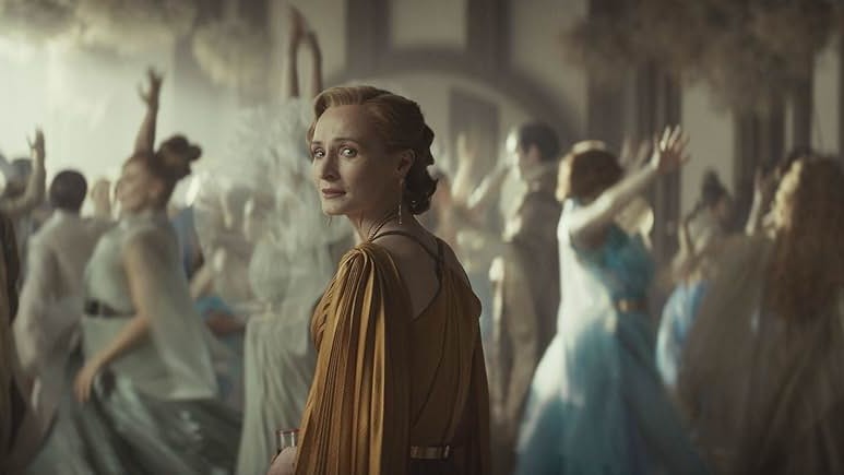 Still from the Star Wars TV show, Andor. A worried Mon Mothma is looking over her shoulder. She is wearing a formal gold gown and is surrounded by lots of dancing people celebrating a wedding.