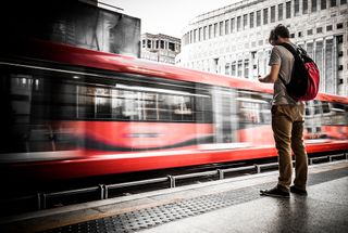 A man stood on a train platform looking at his phone while a blurred red train passes in front