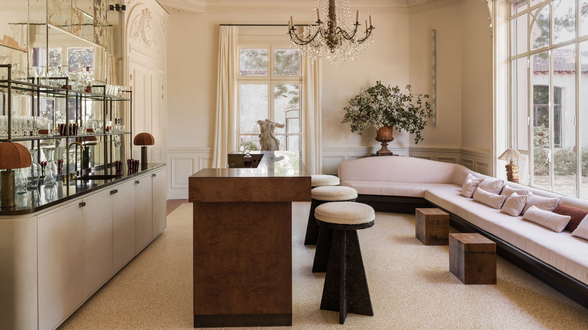 A living room with a long sofa, wood block side tables, a bar with aged mirror details, a wood island, black and white stools, a vase of flowers, and a chandelier