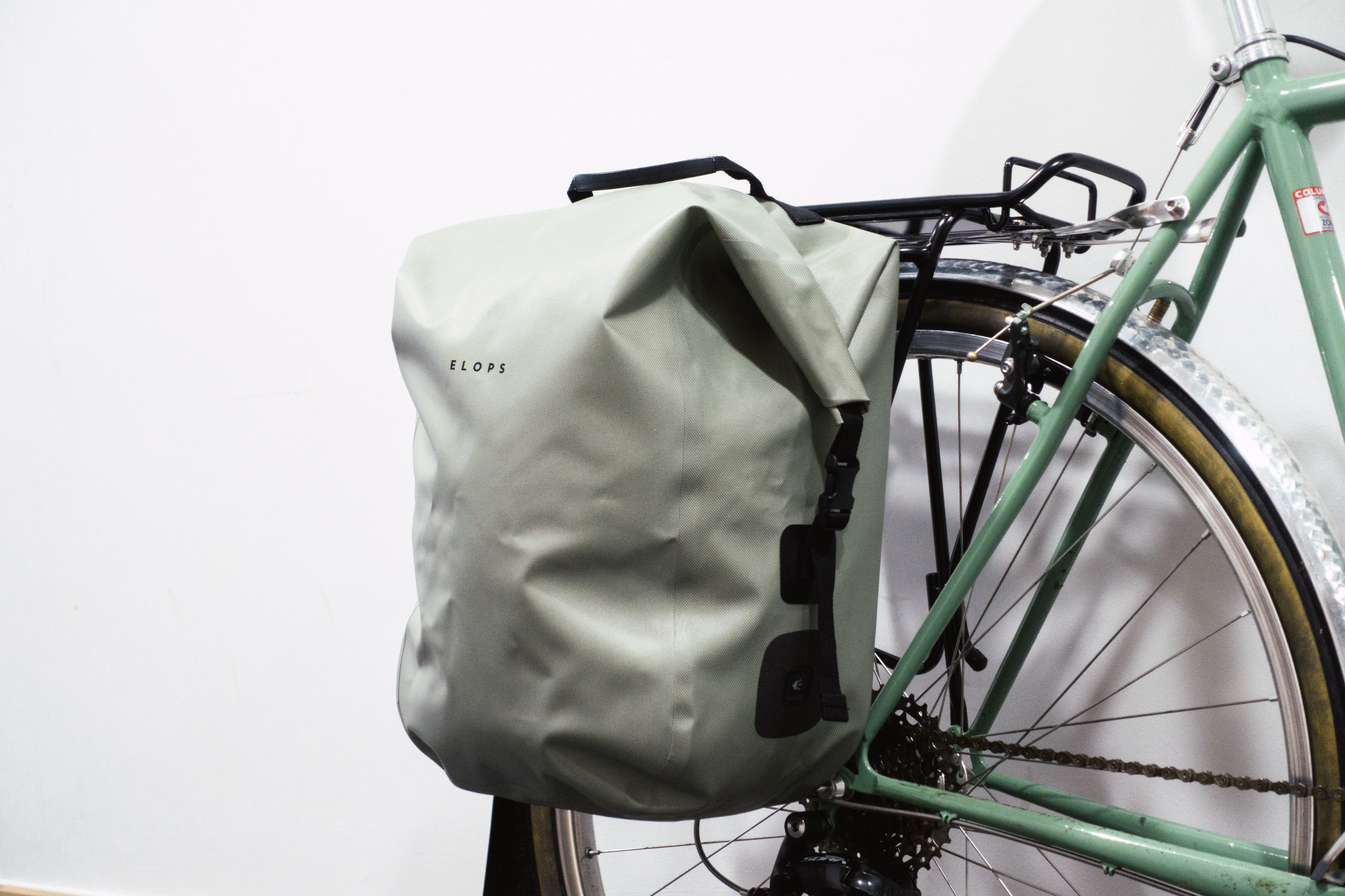 A sage green rolltop pannier mounted to a green bike against a white background