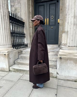 marilyn wears a brown baseball cap, brown long coat, brown reformation patrizia bag, blue jeans and brown loafers.