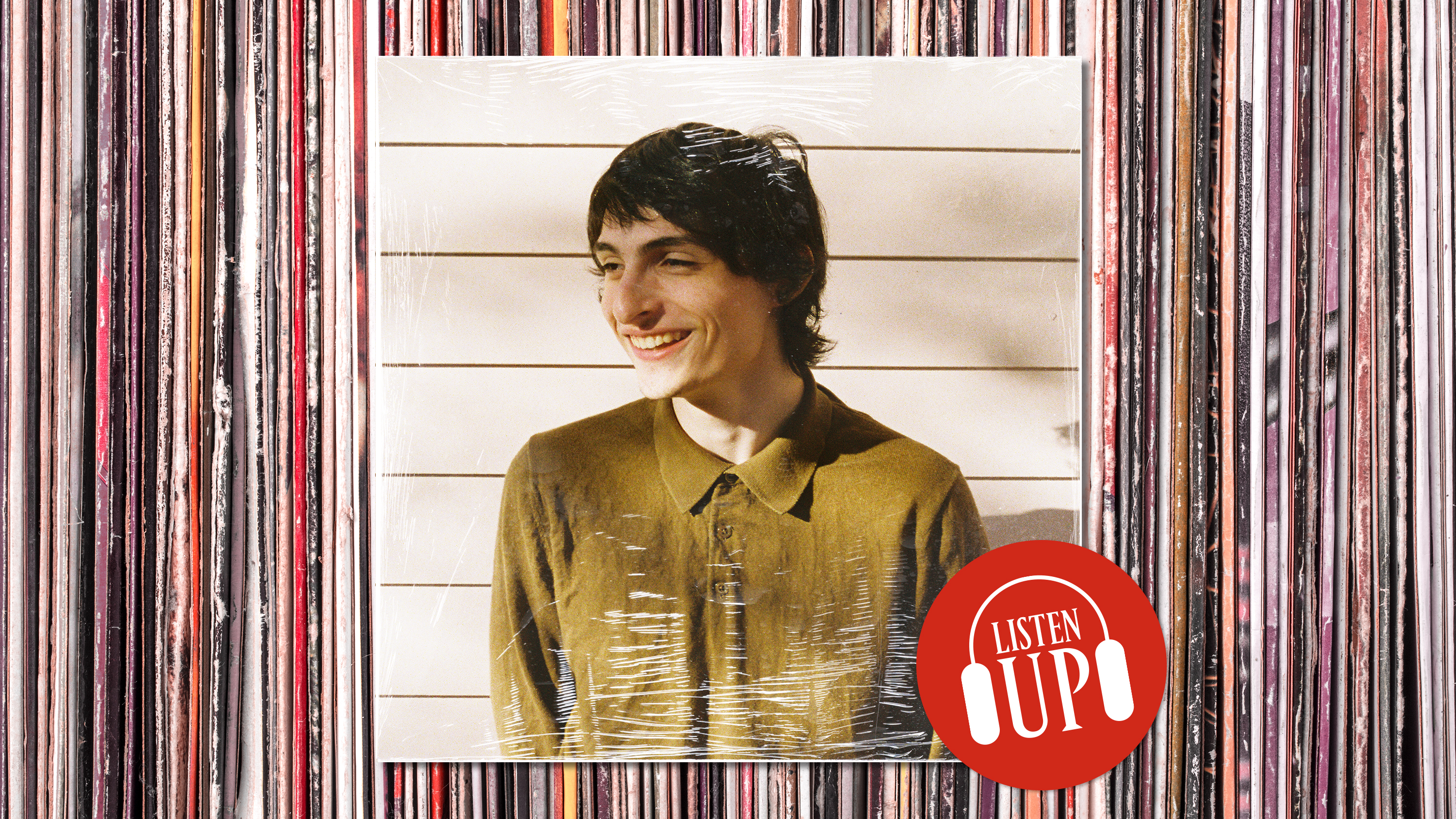 a smiling photo of actor musician finn wolfhard wearing a green button up with a marie claire sticker on it that says listen up and a background that looks like vinyl records