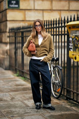 A woman at Milan Fashion Week wearing blue jeans, a white tee, a khaki cardigan sweater, and holding a brown bag.