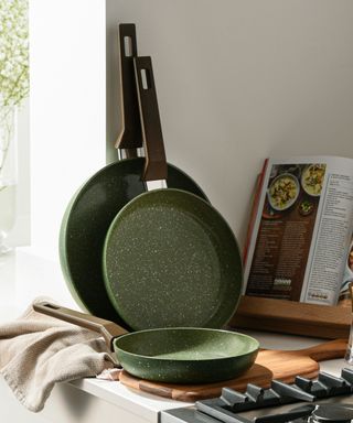 Three dark green pans, one on a wooden cutting board and two leaning against a white wall, with a cooking book open to the right of them and a black hob in front of them