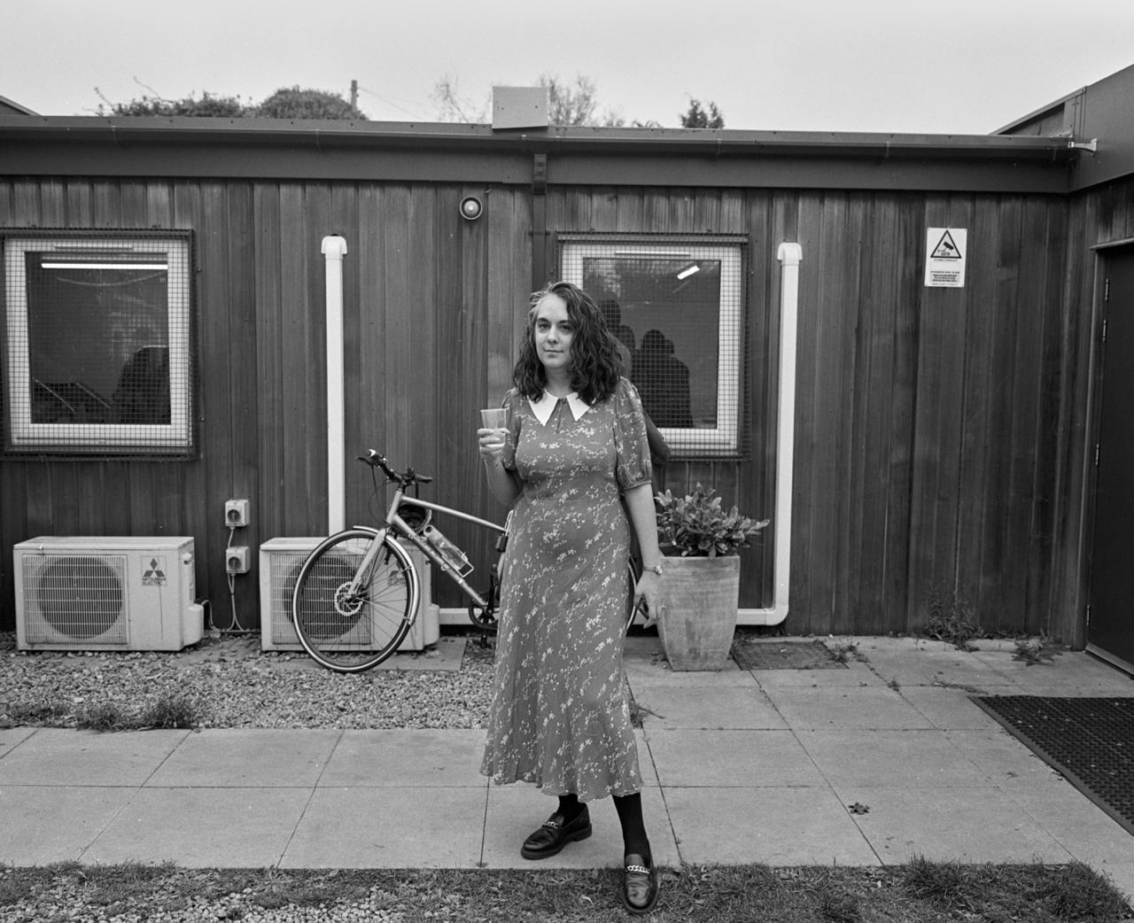 A black-and-white shot of a woman in a long dress with a white, wide collar holding a cup in front of a wooden cabin.