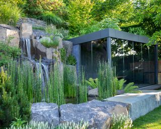 grasses and trees planted around natural stone waterfall beside glass garden building in a sloping garden