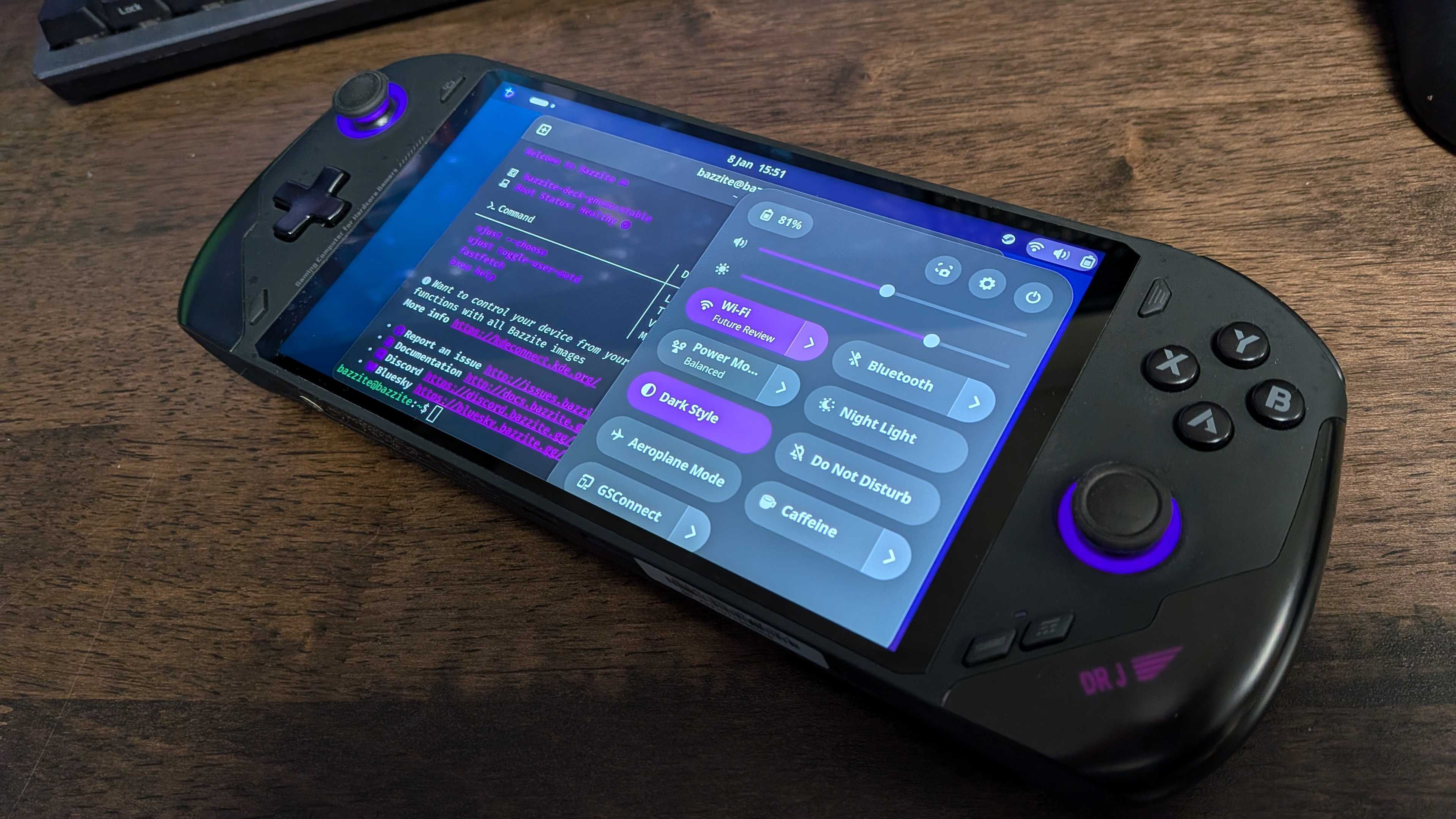 A handheld gaming PC on a desk running Bazzite, a Linux OS, on-screen.