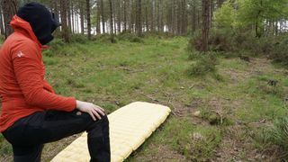 A man in a red jacket turned away from the camera inflates a yellow Forclaz Inflatable Trekking Mattress MT900 in a grassy area in some woodland.