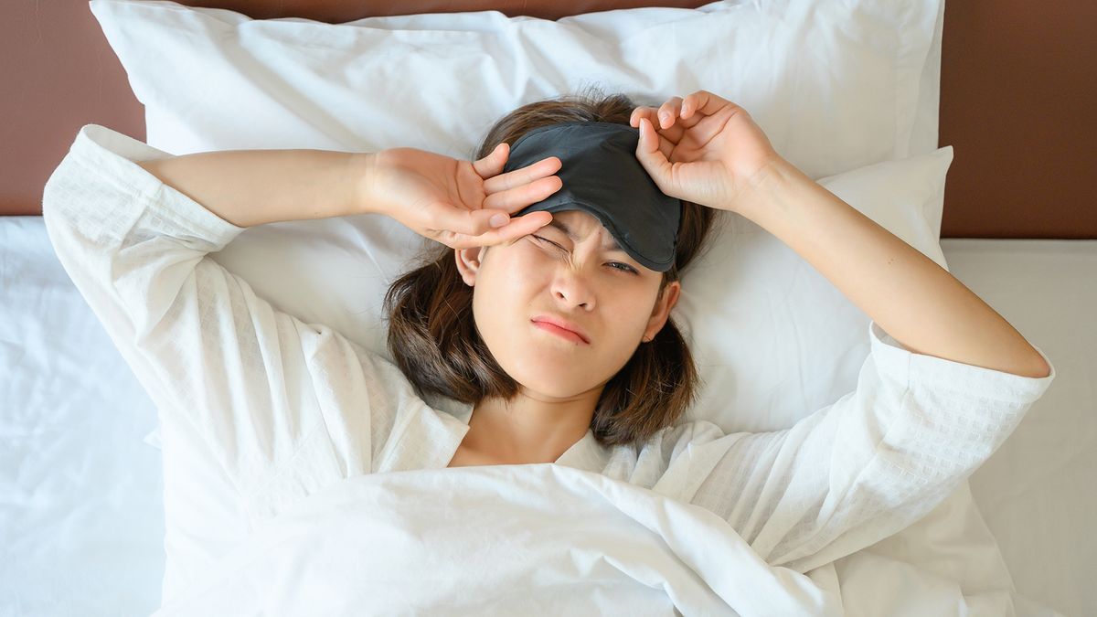 A woman lying on her back wearing an eye mask wakes up after a bad night&#039;s sleep on an unsupportive mattress