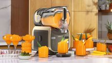 The black and gold Ninja Slushi Max machine filled with orange slush, with several glasses of orange slush surrounding it on a marble counter. 
