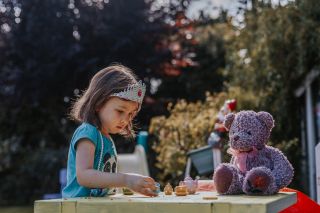 A young girl with a tiara on having a pretend tea party outdoors with her teddy bear.