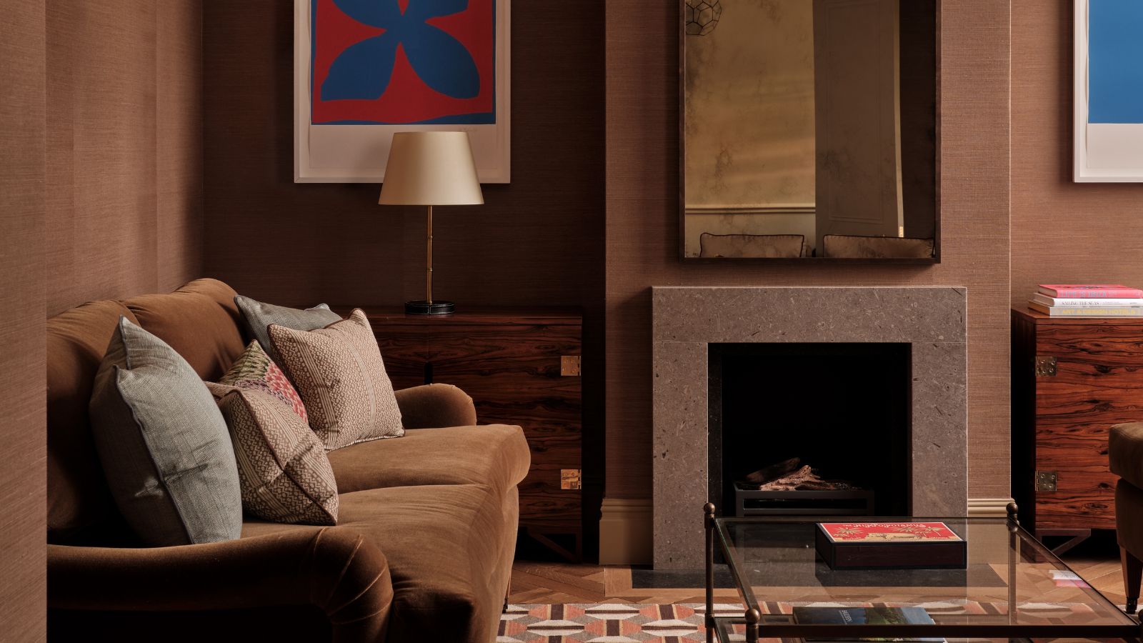 Living room with brown grasscloth walls, cream ceiling, lantern pendant light, geometric orange and brown rug, glass coffee table, brown velvet sofa, and a fireplace with tarnished mirror above it