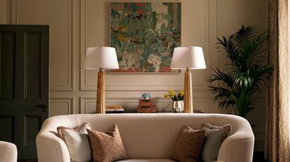A living room with a framed painting, a potted kentia palm, a cream couch with pillows, and behind is a sideboard with a pair of brushed gold and white table lamps, a vase of flowers, and a set of two boxes
