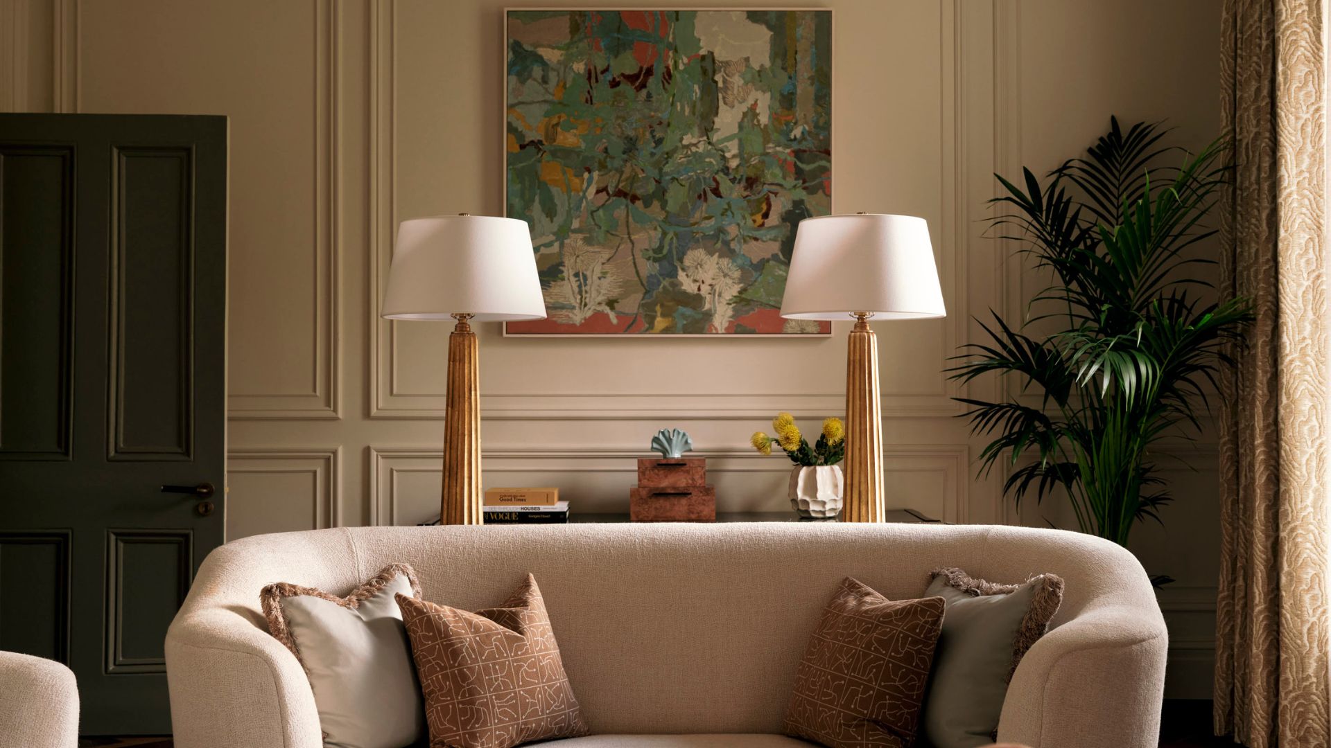 A living room with a framed painting, a potted kentia palm, a cream couch with pillows, and behind is a sideboard with a pair of brushed gold and white table lamps, a vase of flowers, and a set of two boxes