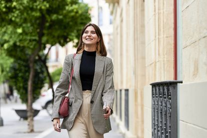 A woman walking on the street, wearing a black top with a grey, patterned blazer, cream trousers, and a red handbag. 