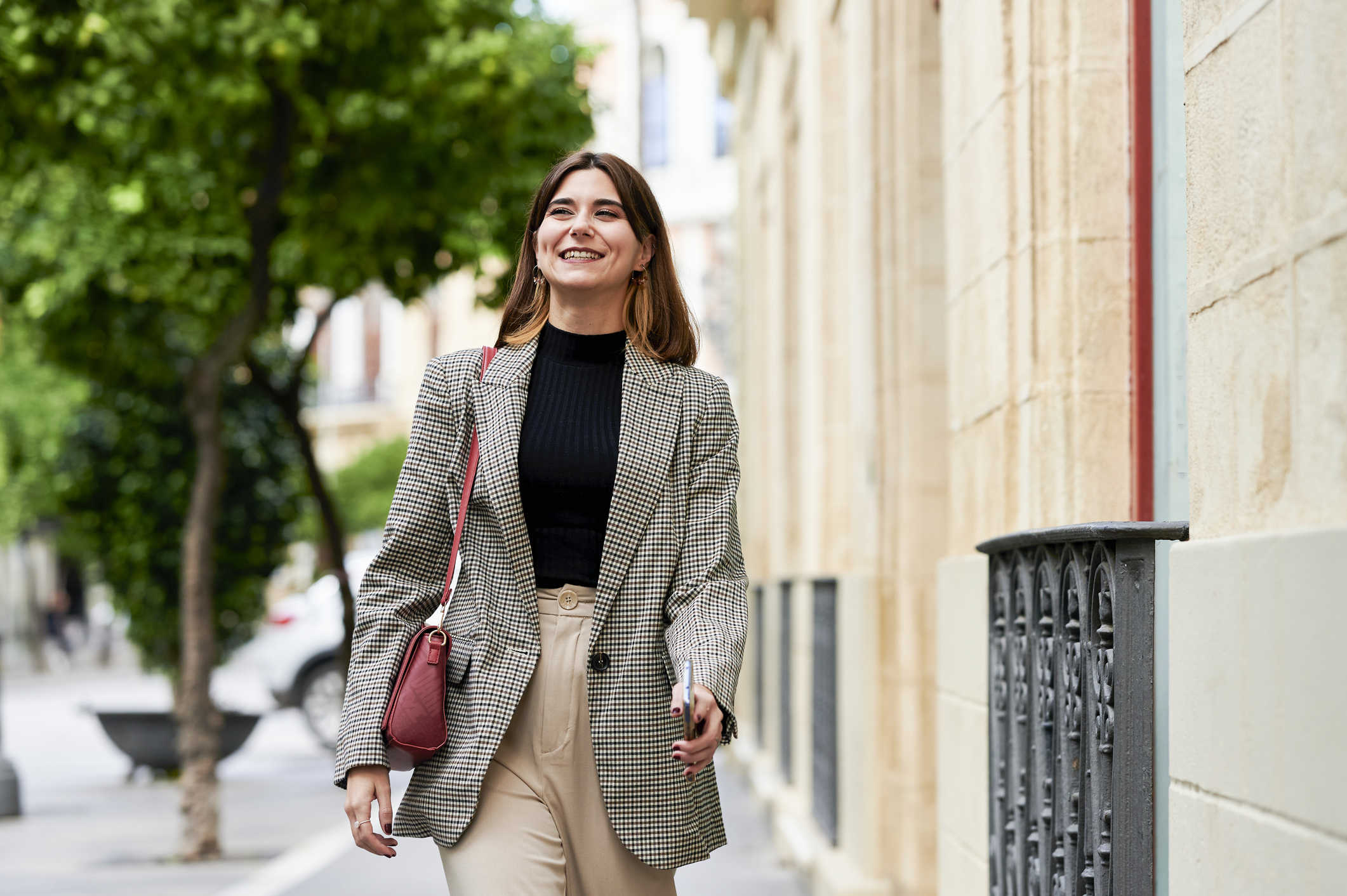 A woman walking on the street, wearing a black top with a grey, patterned blazer, cream trousers, and a red handbag. 