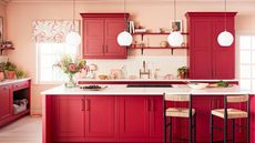 There are no outdated kitchen cabinet colors in this beautiful shot of a kitchen with cherry red cabinets and shelves, two white circular pendant nights, a kitchen island with a hob, a white counter, and two rattan bar chairs