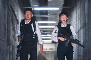 a man and a woman holding guns and wearing bullet vests in a hallway in the action-comedy movie 'Mission Cross.'