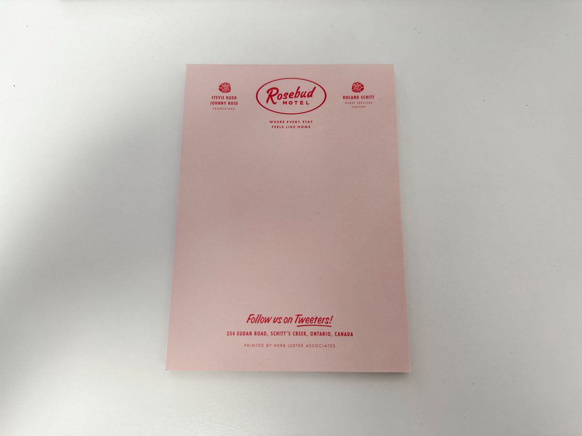 An A5 size pink notepad on a white desk. The notepad has small print Schitt's Creek themed letterheading at the top in red