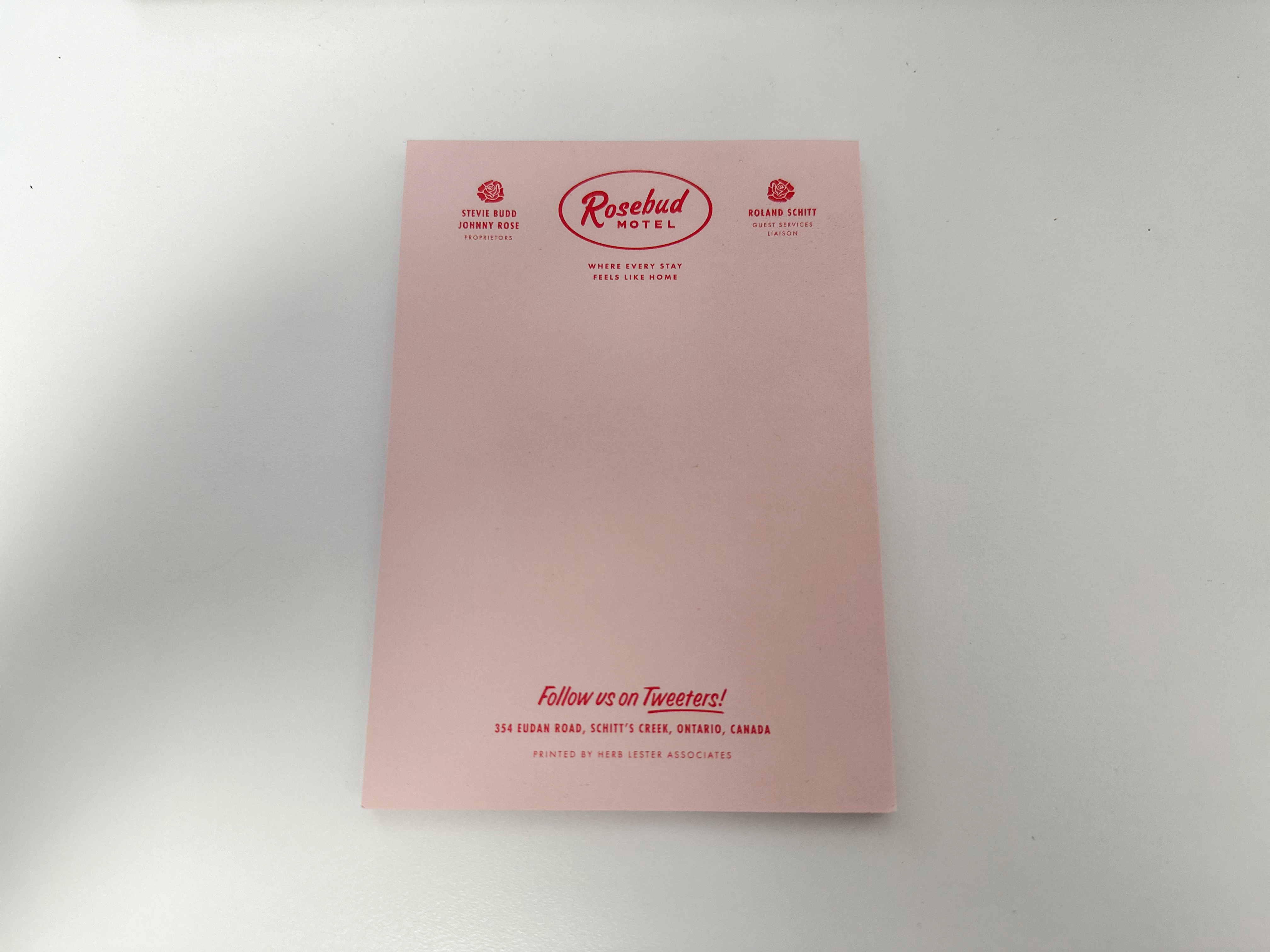 An A5 size pink notepad on a white desk. The notepad has small print Schitt's Creek themed letterheading at the top in red