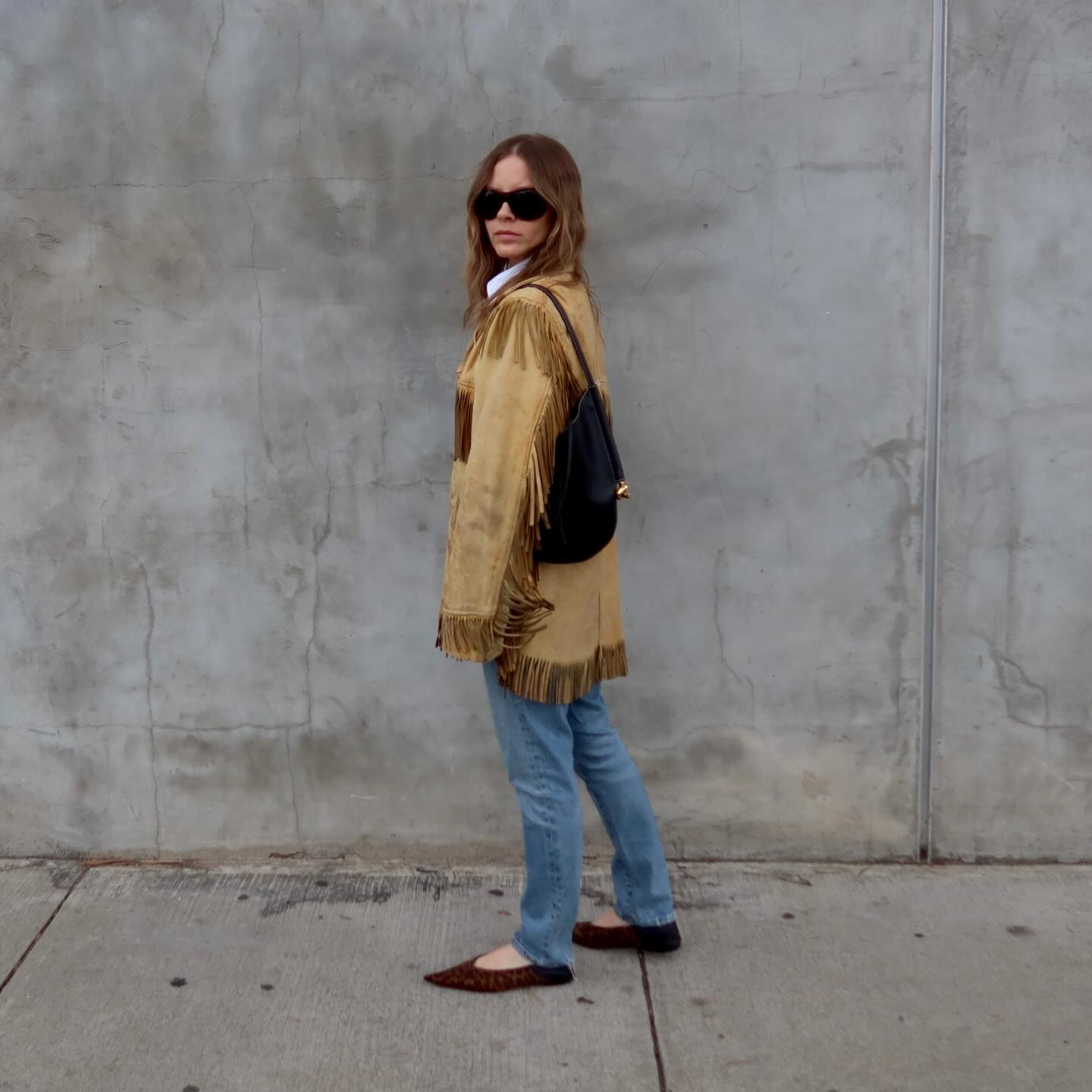 Woman wearing oversize sunglasses, a suede jacket and blue jeans.