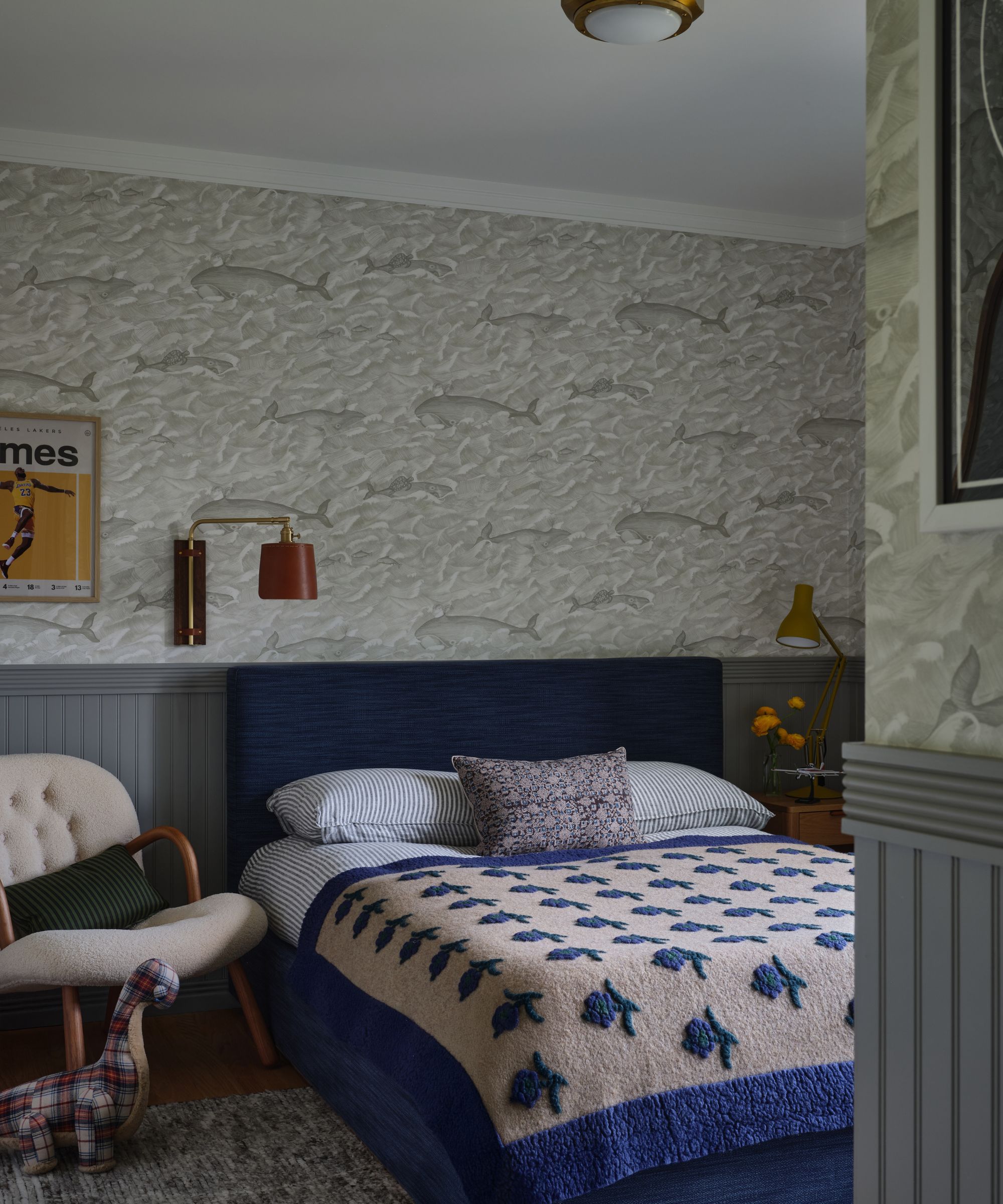 Child&#039;s bedroom featuring a navy upholstered headboard, a blue and floral patterned quilt, a beige whale-patterned wallpaper, and grey wainscoting