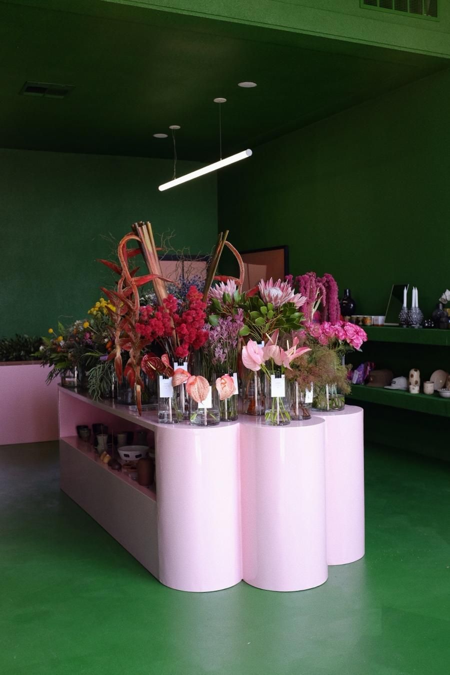 A color-blocked flower shop with green walls and floors and pink flower displays is brought to life by natural as well as neon lights and the many colorful specimen sitting in its glass vases.