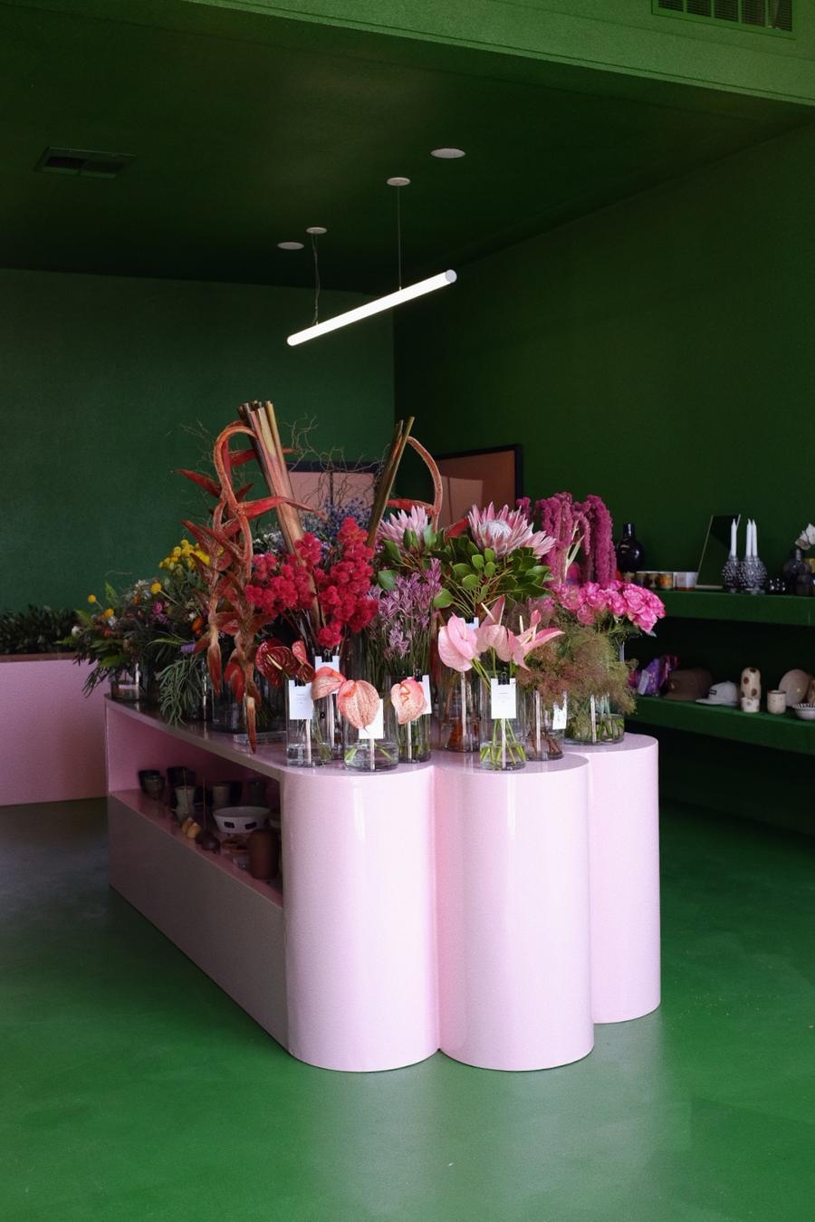 A color-blocked flower shop with green walls and floors and pink flower displays is brought to life by natural as well as neon lights and the many colorful specimen sitting in its glass vases.