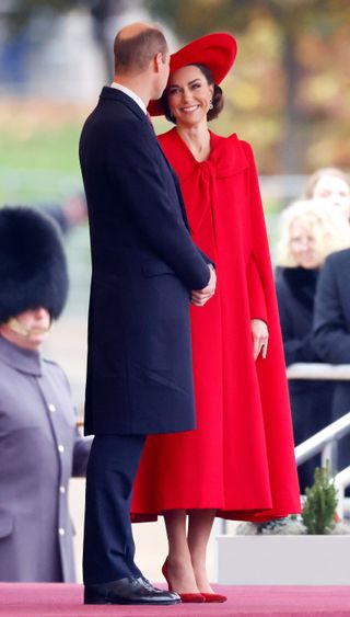 Kate Middleton wearing a red cape and hat smiling at Prince William