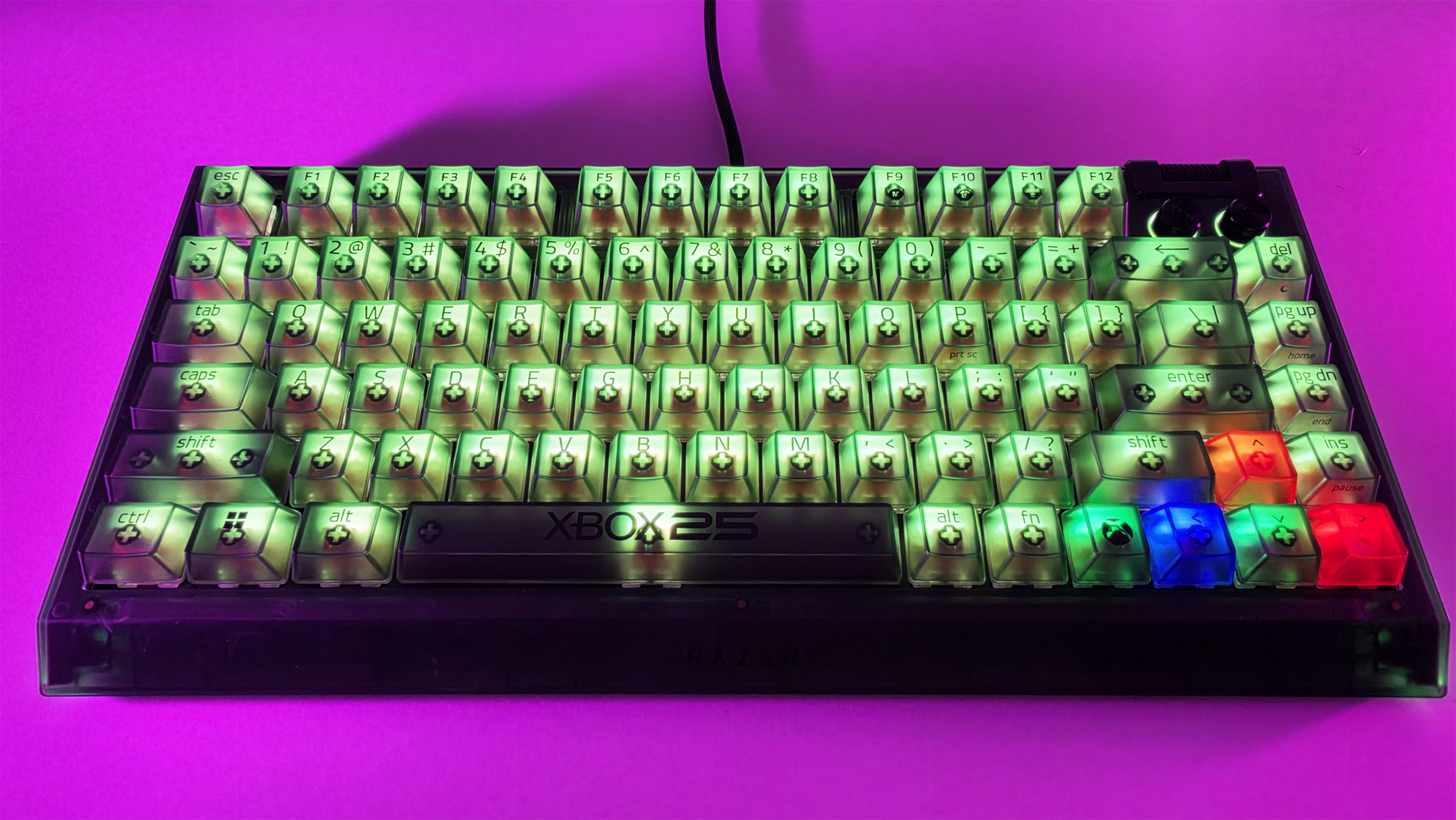 Front view of the Razer BlackWidow V4 75% Xbox 25th Anniversary mechanical keyboard lit with green and multicolored RGB on a pink background.