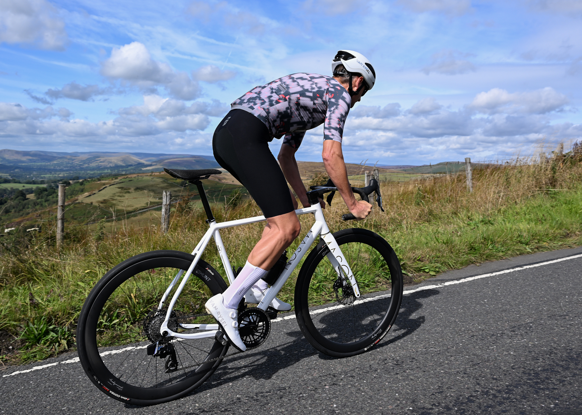 a white road bike being ridden out of the saddle uphill by a man wearing white shoes, white helmet, black shorts and a patterned grey jersey with countryside hills in the background