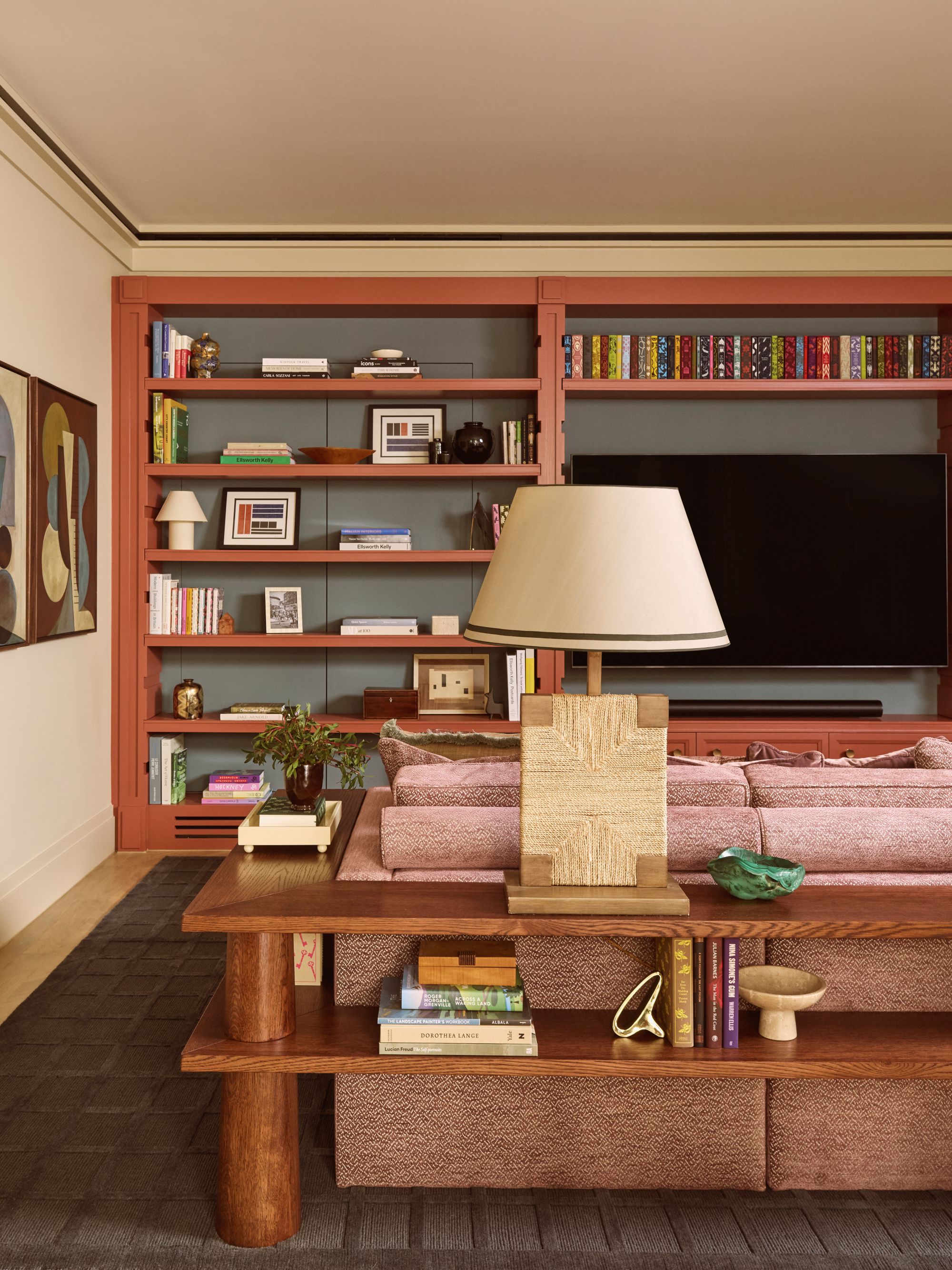 a media room with a large built-in bookcase filled with books, picture frames, and decorative objects. In the foreground is a custom shelf framing a clay red sofa, holding more books, objects, and a table lamp