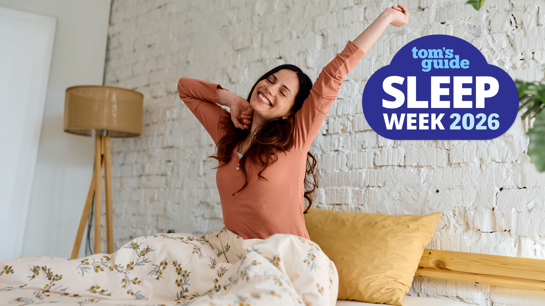 A woman with long dark brown hair wearing an orange night shirt wakes up happy in a bright and light bedroom, with her arms outstretched, and smiling because she had a great night's sleep and now feels full of energy and vitality for the day ahead. A blue Tom's Guide Sleep Week 2026 logo sits in the top right hand corner