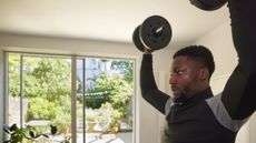 Man at home wearing long sleeve top lifting dumbbells up overhead