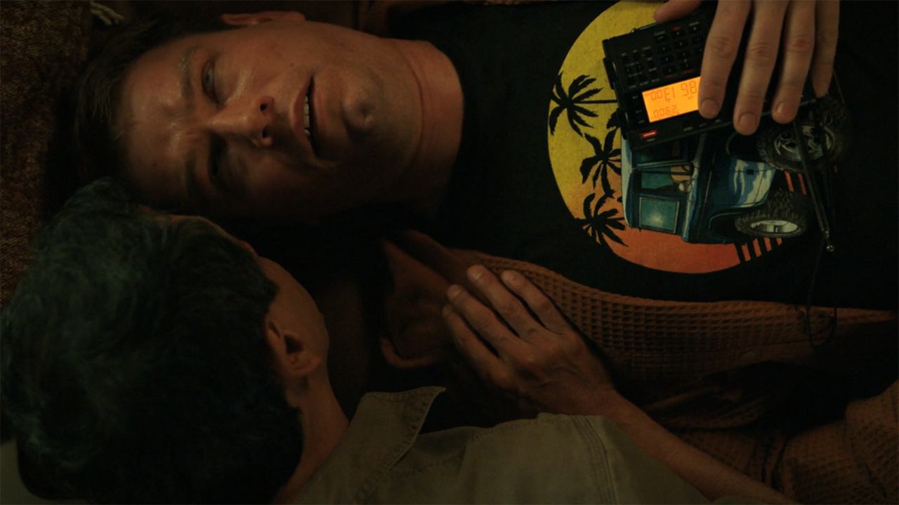 Manousos speaking to a convulsing person with a portable radio lying on them in Pluribus episode 9