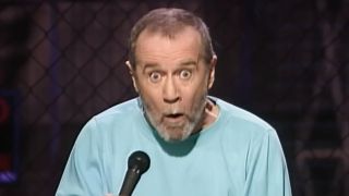 George Carlin performing, in a blue shirt, making a funny 'oh no' face, archival footage for HBO's doc george Carlin's American Dream.