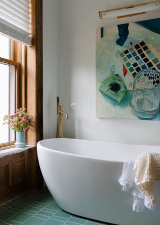 freestanding bath tub by a wooden window, blue tiled beneath and a green-blue oil painting hangs above the bath